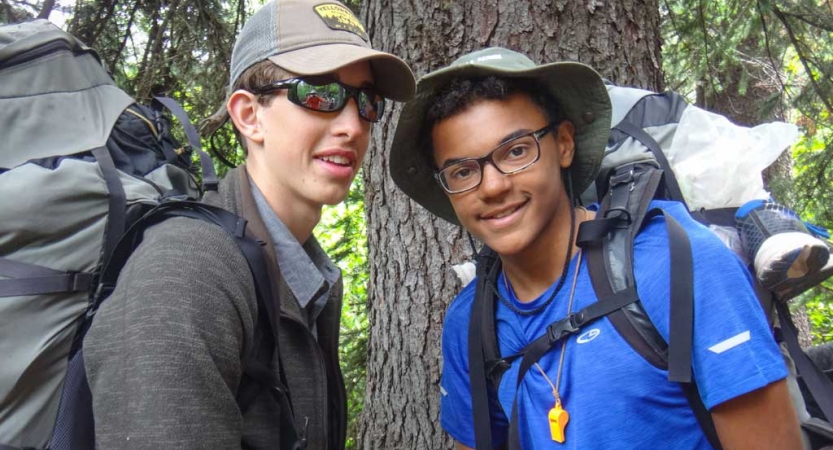 Two students wearing backpacks smile at the camera in front of a tree. 